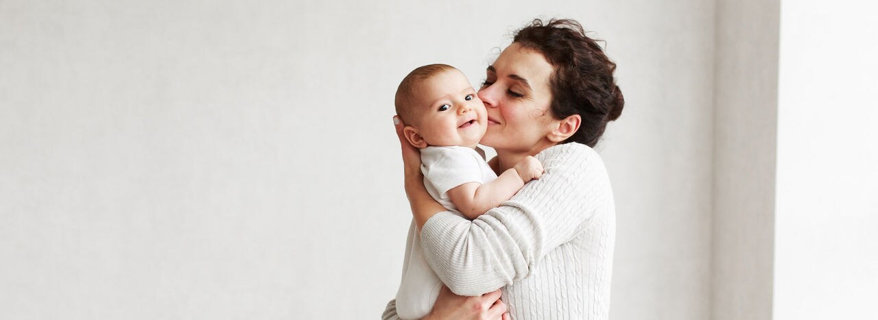 Beautiful caucasian middleaged woman with dark hair in home cozy clothes at home by the window holding a three-month-old baby on a white wall background